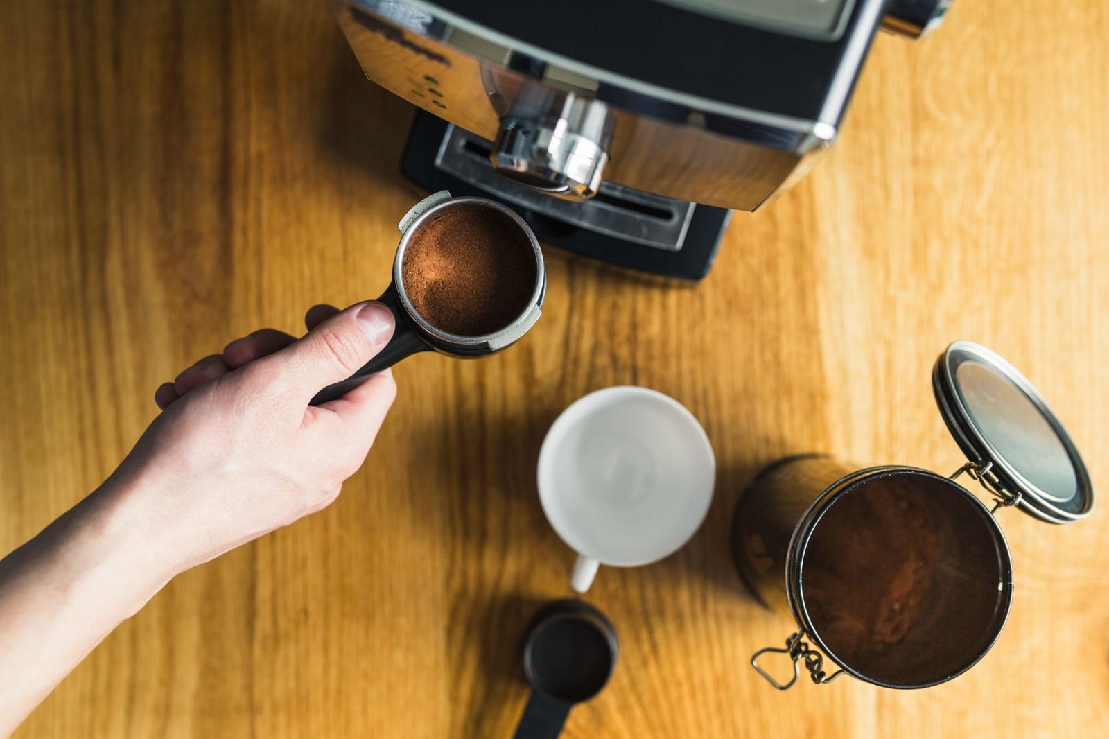 A hand holding a portafilter of fresh grounds beside a grinder